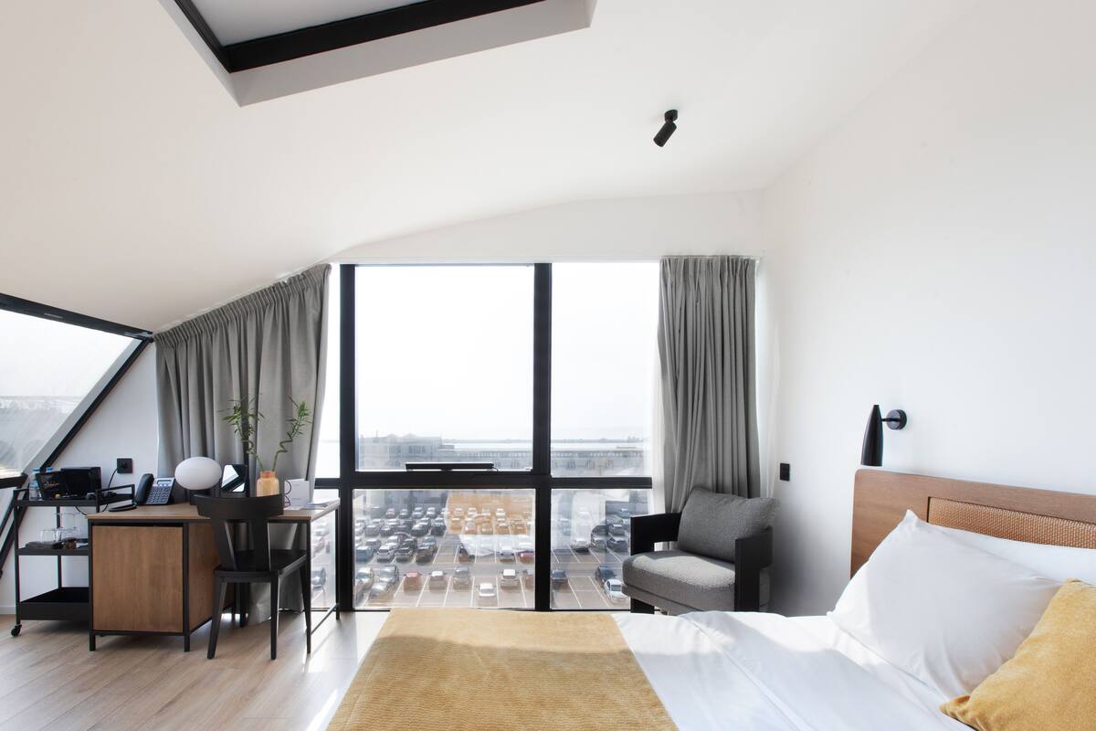 A modern hotel room with a large window, a desk with a computer, a comfortable bed, and a view of the city.
