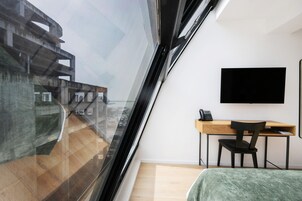 Una camera moderna con una grande finestra, un letto, una scrivania con una sedia e una TV a schermo piatto fissata al muro.