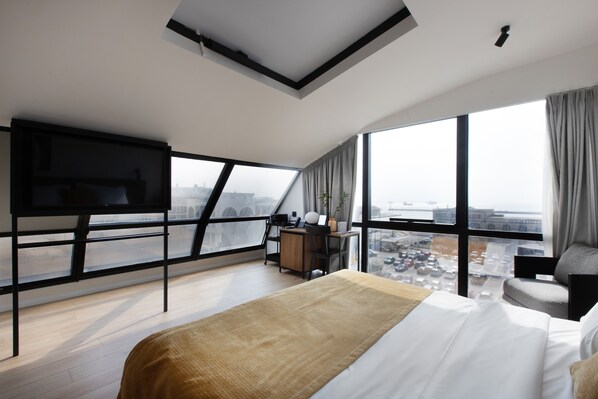 A modern hotel room with a large bed, a desk, and a TV. The room has large windows with a city view. - Noa Hotel (Thessaloniki)