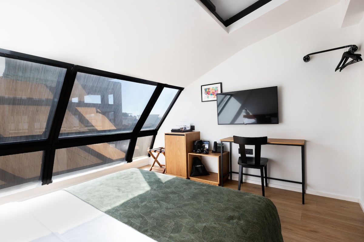 A modern bedroom with a large window, a bed, a desk, and a TV.