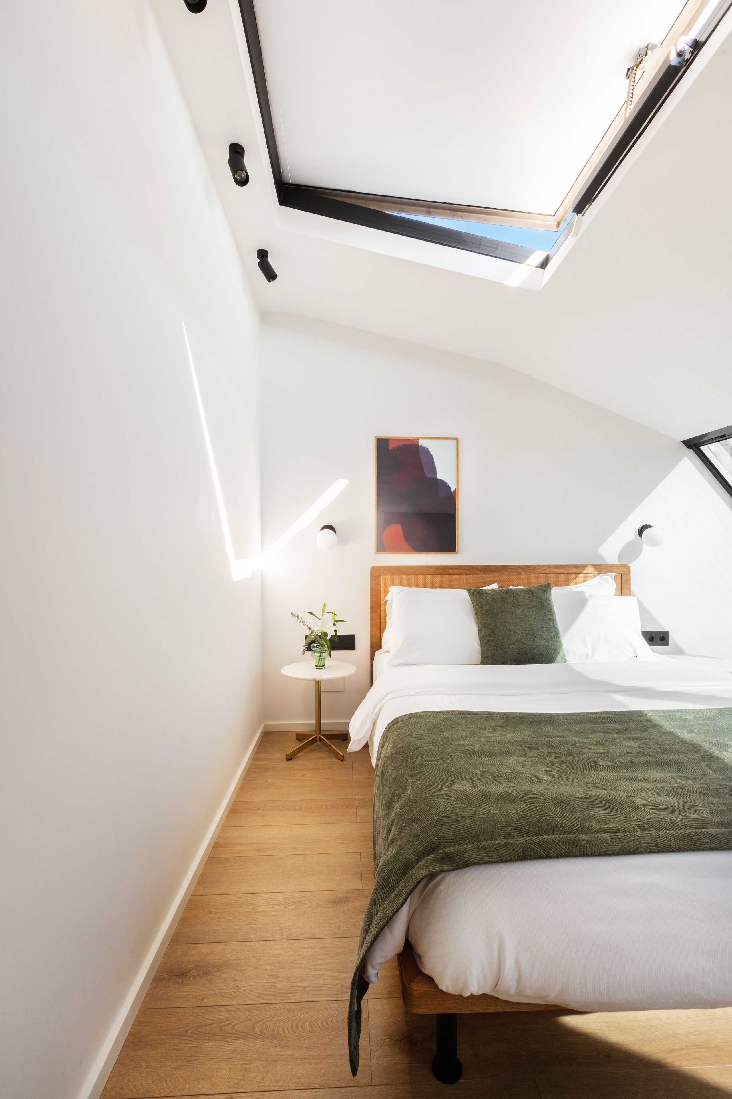 A modern bedroom with a skylight, a wooden bed, a small bedside table with a vase, and a wall-mounted abstract painting.