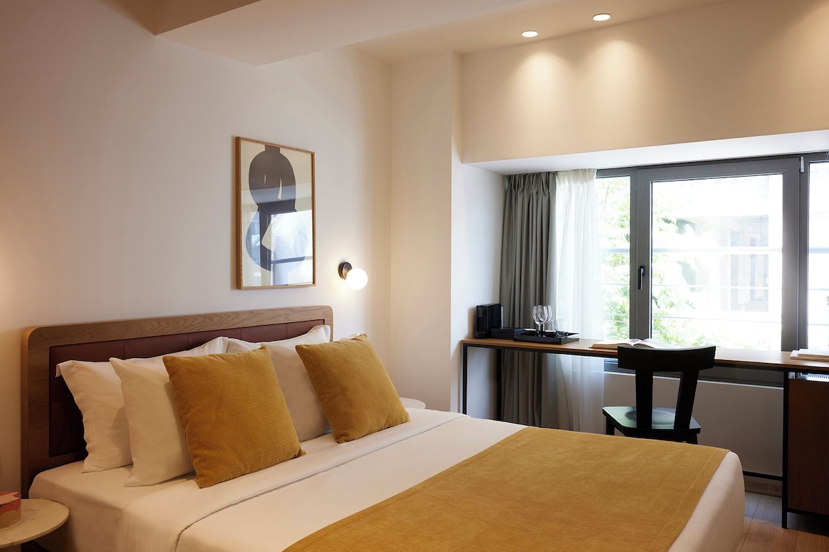 A hotel room with a bed, a desk, a chair, a window with curtains, and a wall-mounted light fixture.