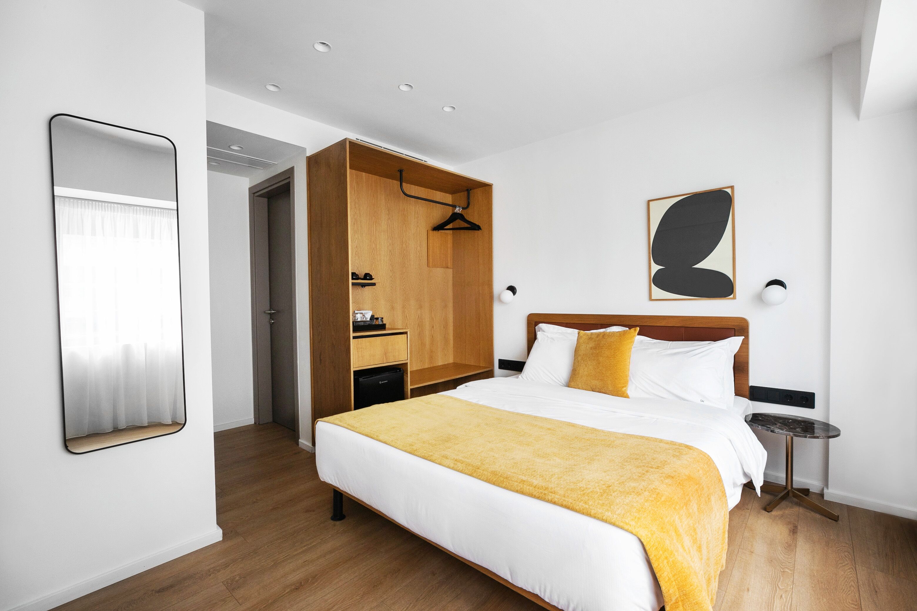 A hotel room with a large bed, a wooden wardrobe, a bedside table, and a wall-mounted artwork.