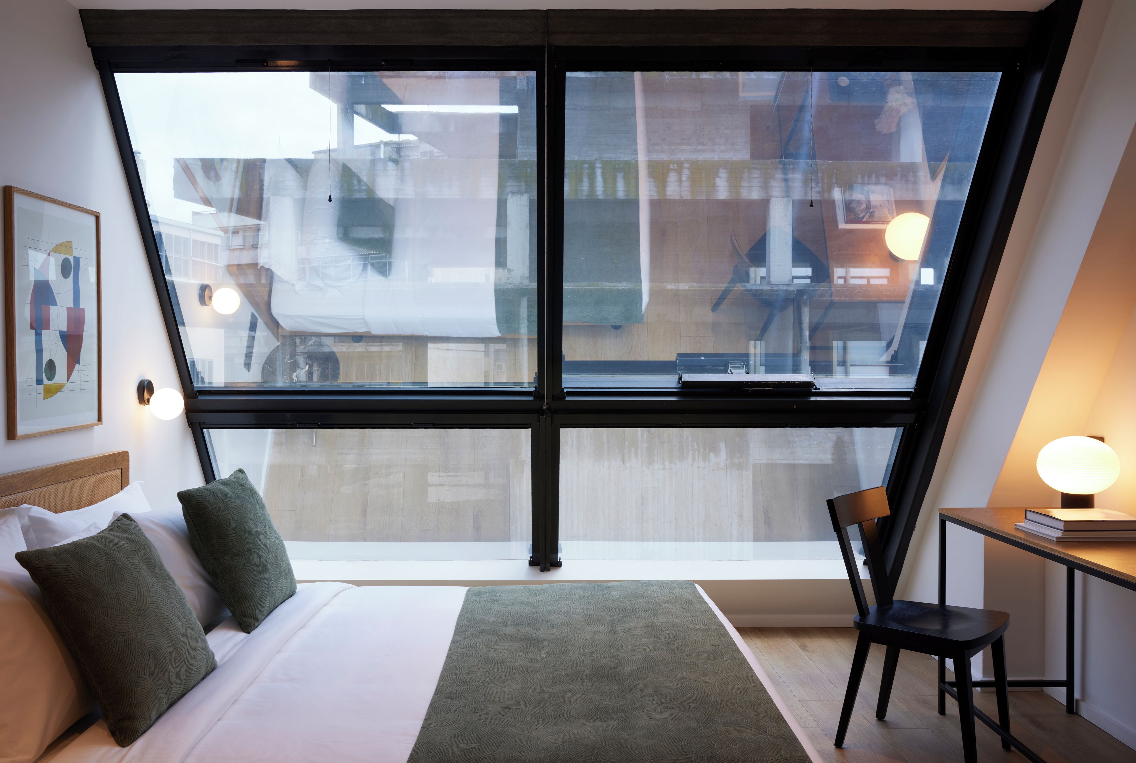 Ein modernes Schlafzimmer mit großem Fenster, einem Bett mit Kissen, einem Schreibtisch mit Stuhl und einer Lampe.