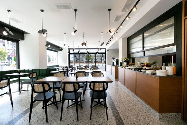 A modern restaurant interior with high tables, chairs, and a counter with food items. - Noa Hotel (Thessaloniki)