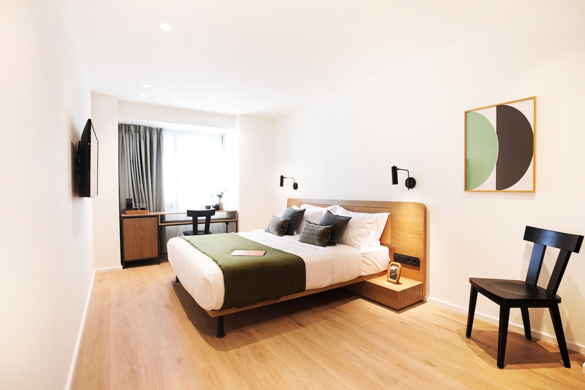 A modern hotel room with a wooden bed, a desk, a chair, and a large window with curtains.