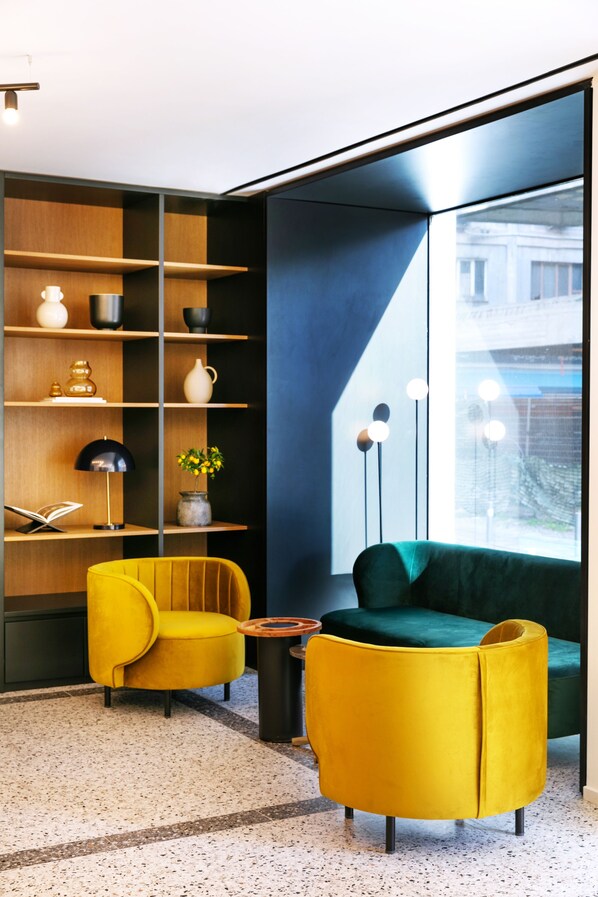 A modern living room with a yellow armchair, a green sofa, a wooden bookshelf, and a large window with a city view. - Noa Hotel (Thessaloniki)