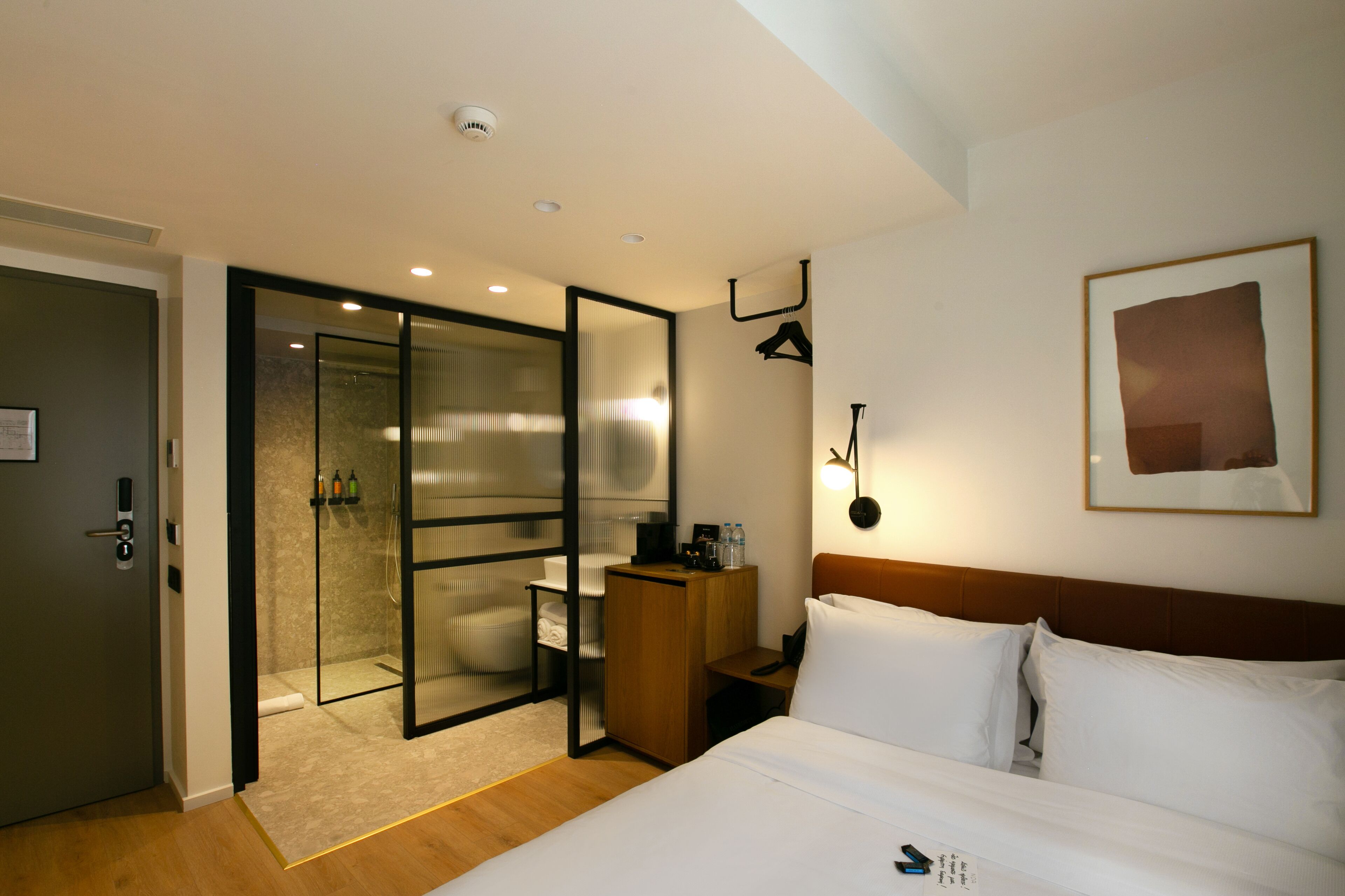 Une chambre d’hôtel moderne dotée d’un grand lit, d’un bureau, d’une douche séparée par des parois en verre et d’un tableau encadré au mur.
