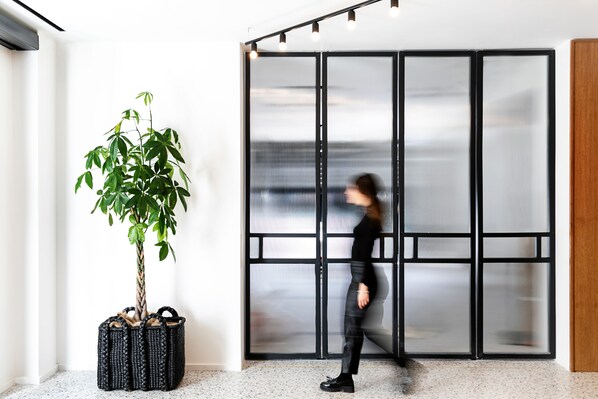 A modern interior with a large potted plant, a sliding glass door, and a person walking by. - Noa Hotel (Thessaloniki)