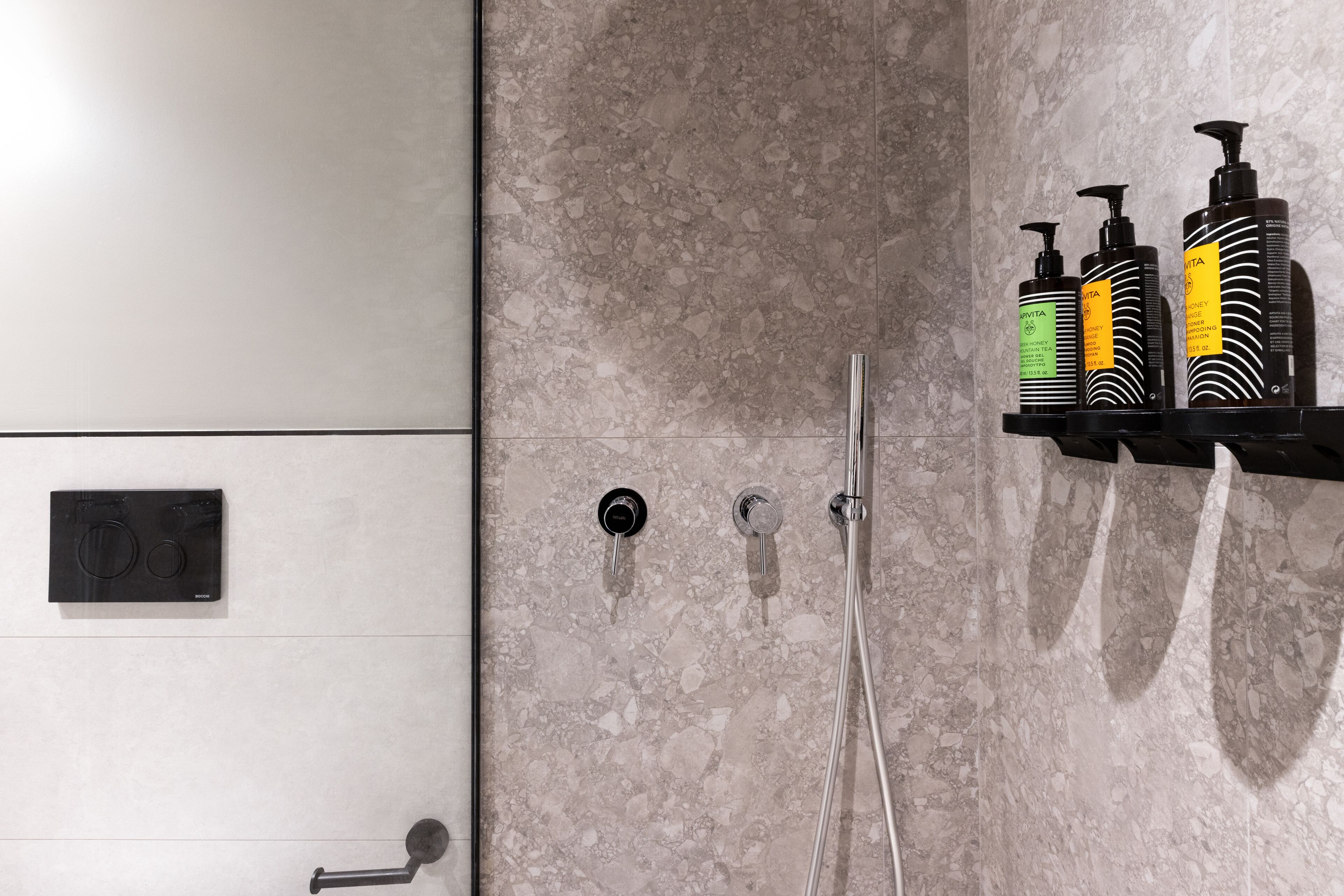A bathroom with a marble wall, a shower with a handheld showerhead, and a shelf with three bottles of shampoo.