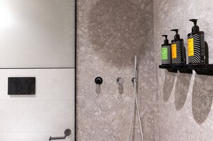 A bathroom with a marble wall, a shower with a handheld showerhead, and a shelf with three bottles of shampoo.
