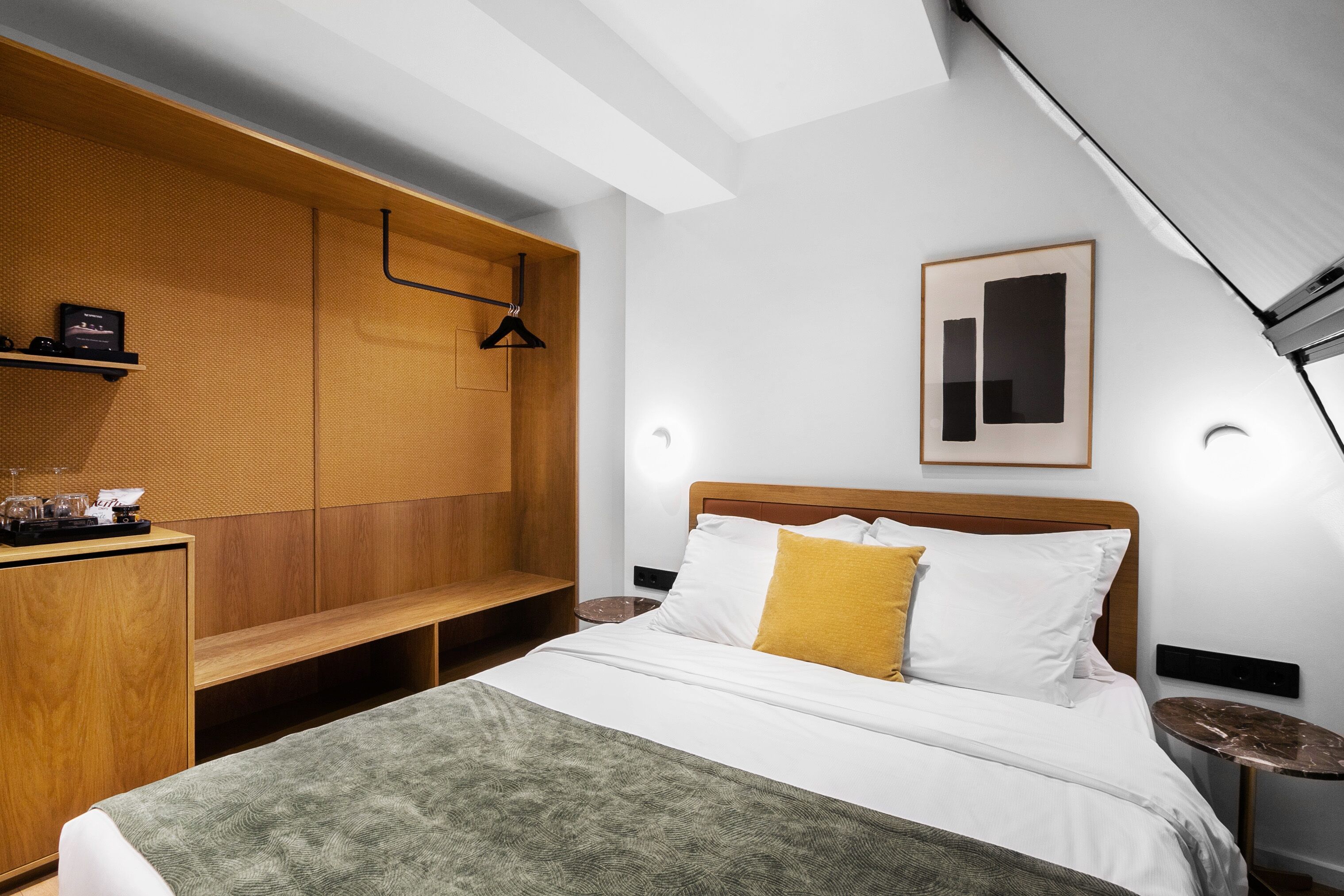 A modern hotel room with a wooden headboard, a bed with white and yellow pillows, a bench, and a small table with a lamp.