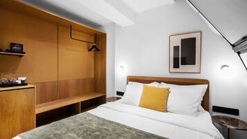 A modern hotel room with a wooden headboard, a bed with white and yellow pillows, a bench, and a small table with a lamp.