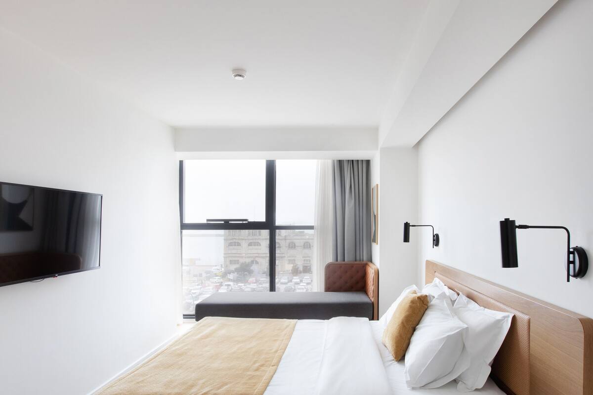 A modern hotel room with a large bed, a TV mounted on the wall, and a city view through the window.
