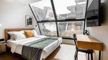 Una habitación de hotel moderna con un ventanal grande, una cama con una manta verde, una mesita de noche de madera y un escritorio con una silla.