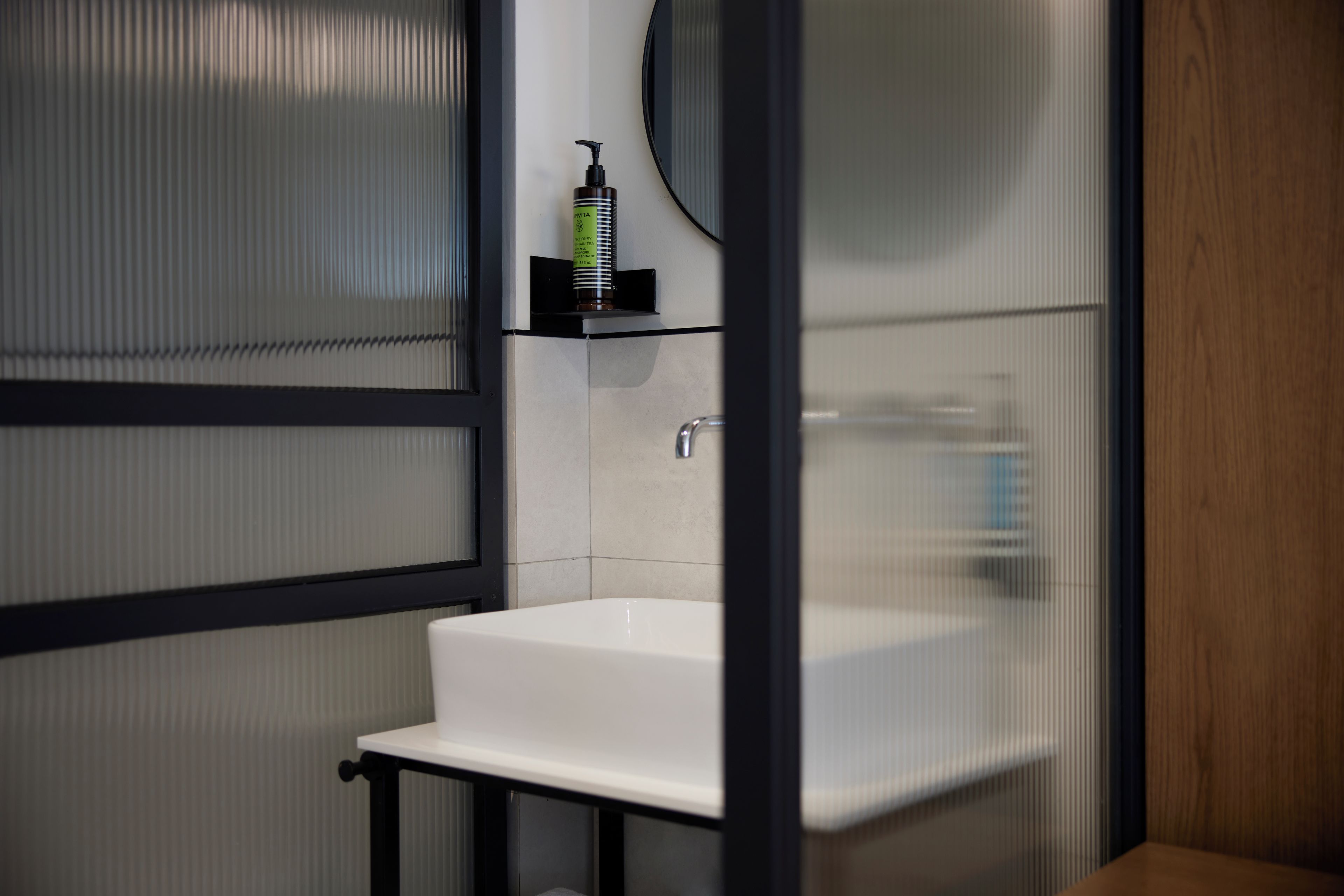 A bathroom with a white sink, a mirror, and a bottle of shampoo on a shelf.