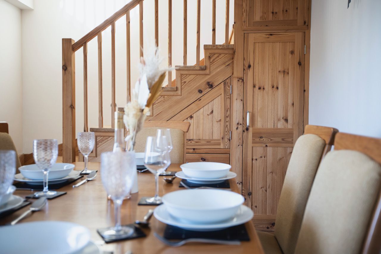 A dining table set for six with white plates, clear glassware, and a vase with white flowers. A wooden staircase with a railing is visible in the background.