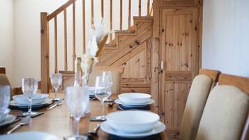 A dining table set for six with white plates, clear glassware, and a vase with white flowers. A wooden staircase with a railing is visible in the background.