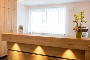 A reception desk with a wooden finish, a vase with orchids, and a window with blinds.