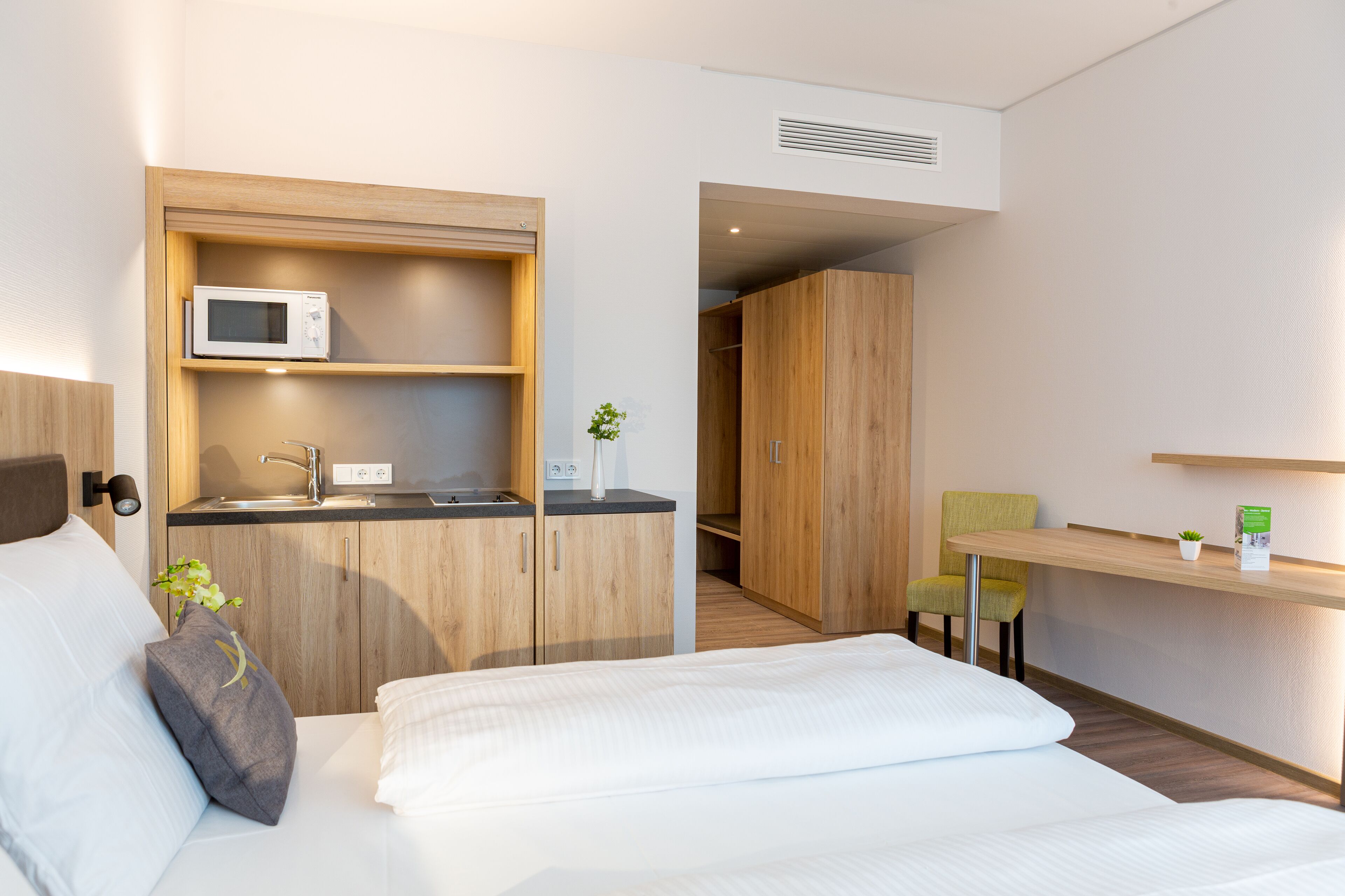 Ein modernes Hotelzimmer mit einem Bett, einer kleinen Küchenzeile, einem Holzschrank, einem Schreibtisch mit Stuhl und einer kleinen Pflanze.