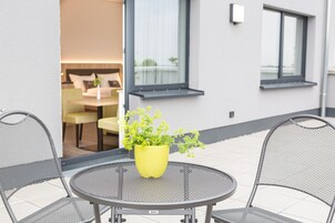 A patio area with a round metal table and two mesh chairs, a yellow potted plant, and a glimpse of an indoor seating area with a sofa and a table.