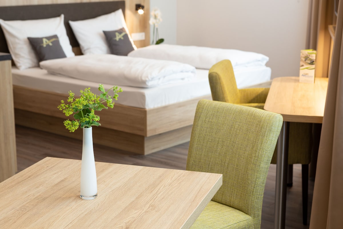 A hotel room with a bed, a small table with a vase, and a green chair.