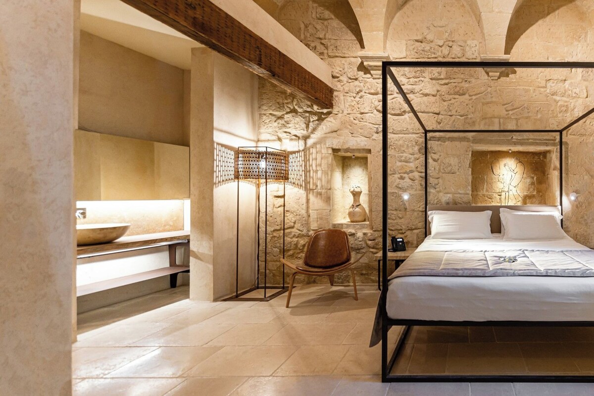 Una cama con dosel en una habitación de paredes de piedra, con un banco de piedra y una mesa pequeña con un jarrón.