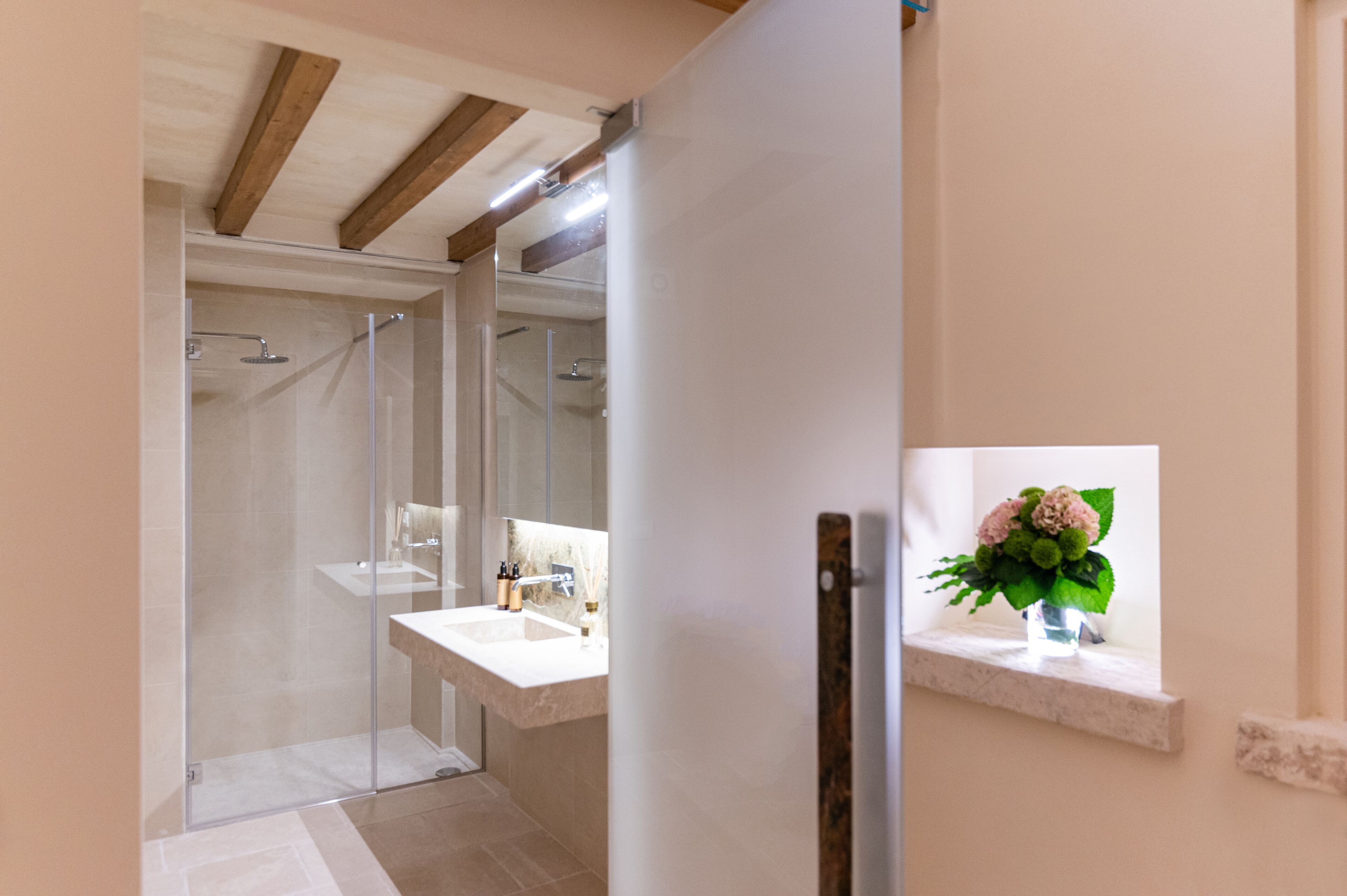 A modern bathroom with a glass-enclosed shower, a white sink, and a small window with a flower vase.