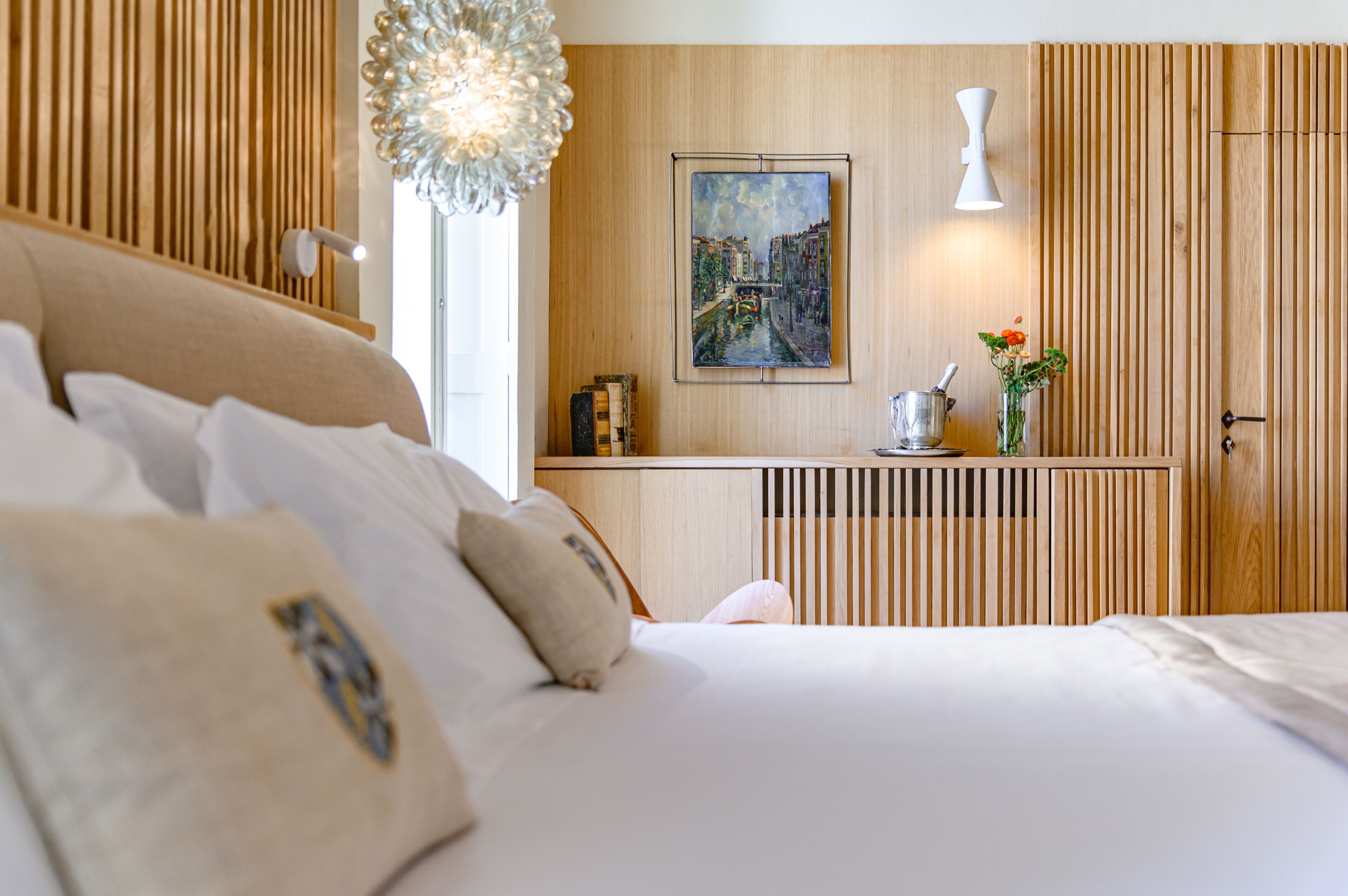 A modern hotel room with a bed, a bedside table with a lamp, a painting on the wall, and a wooden panel design.