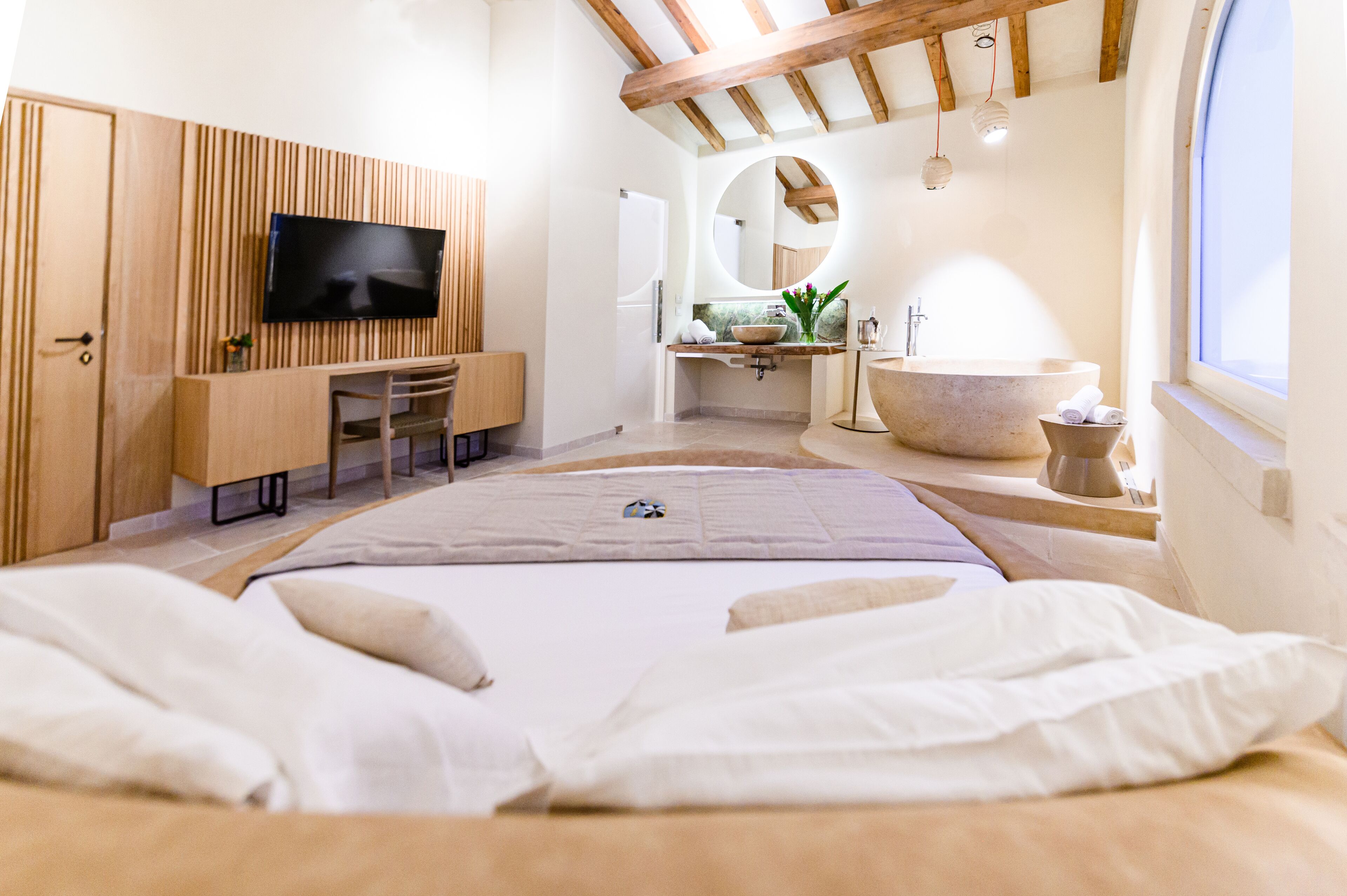 A bedroom with a bed, a desk, a television, a large mirror, a bathtub, and a window.