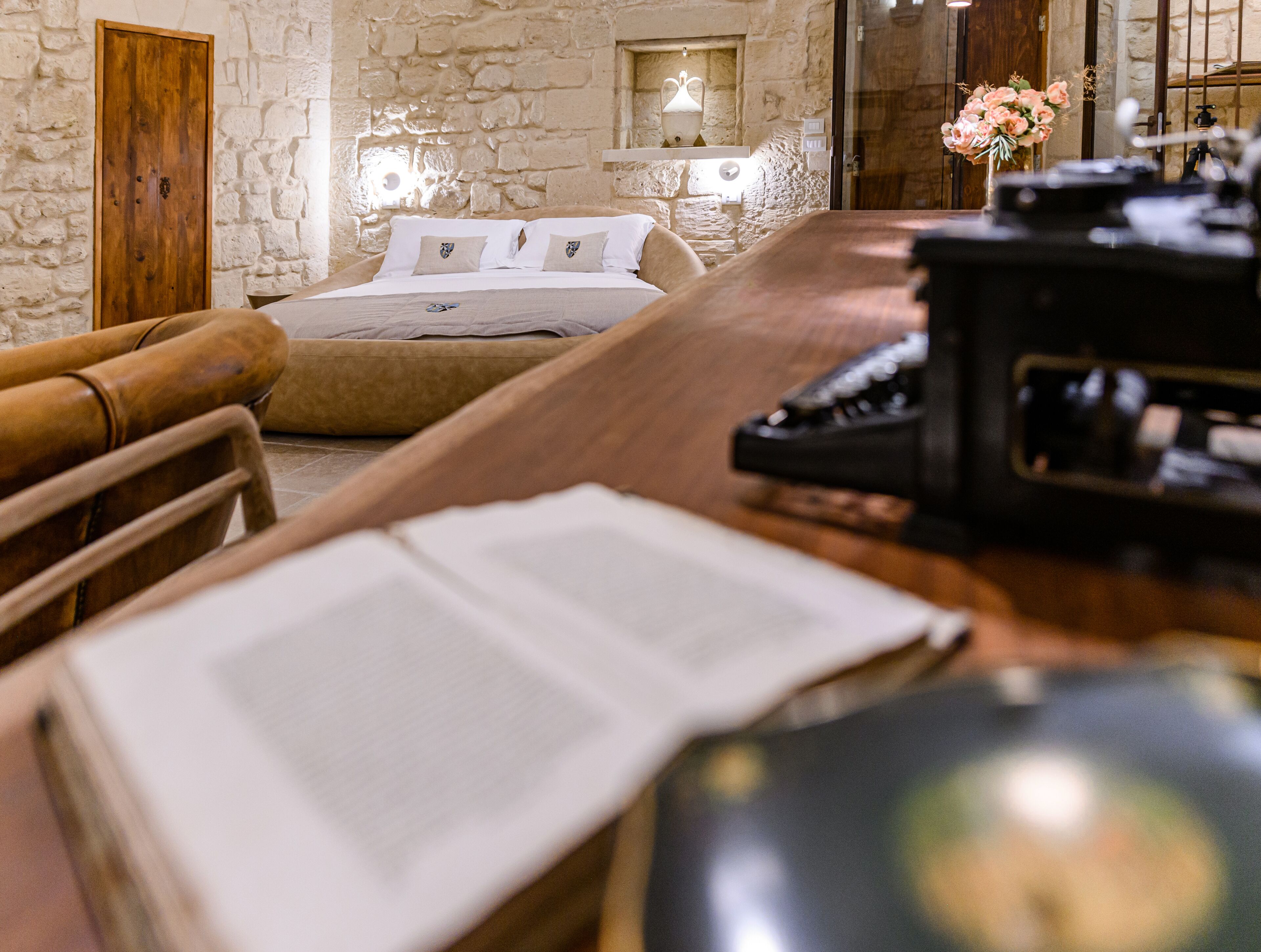 A room with a stone wall, a wooden door, a bed with white bedding, and a desk with a computer.