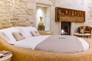 Una cama con manta y almohadas color beige, una pared de piedra, una chimenea con un cuadro encima y una mesita con un florero.