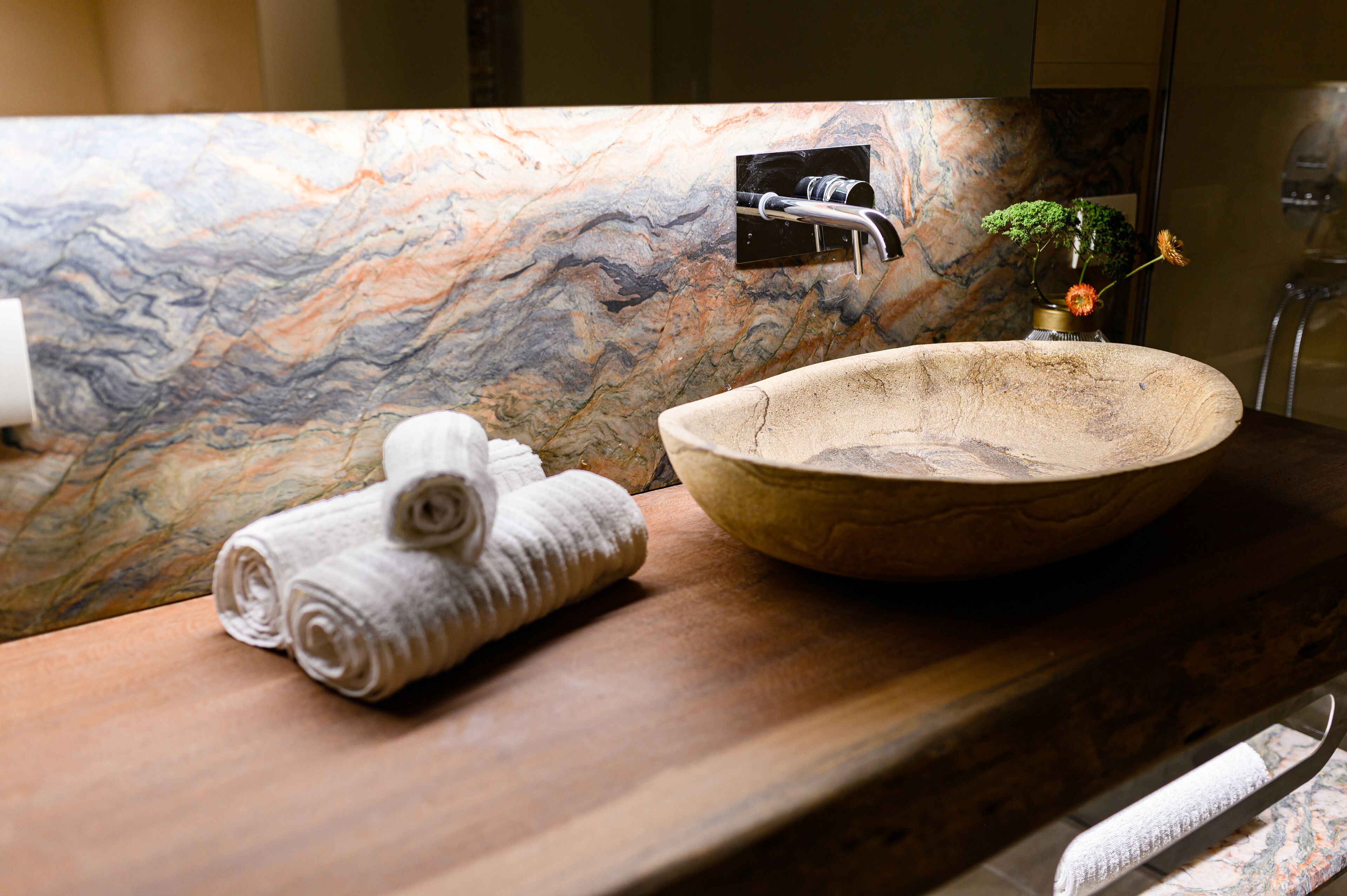 A bathroom with a marble backsplash, a wooden vanity, a large bowl sink, a towel, and a small vase with flowers.