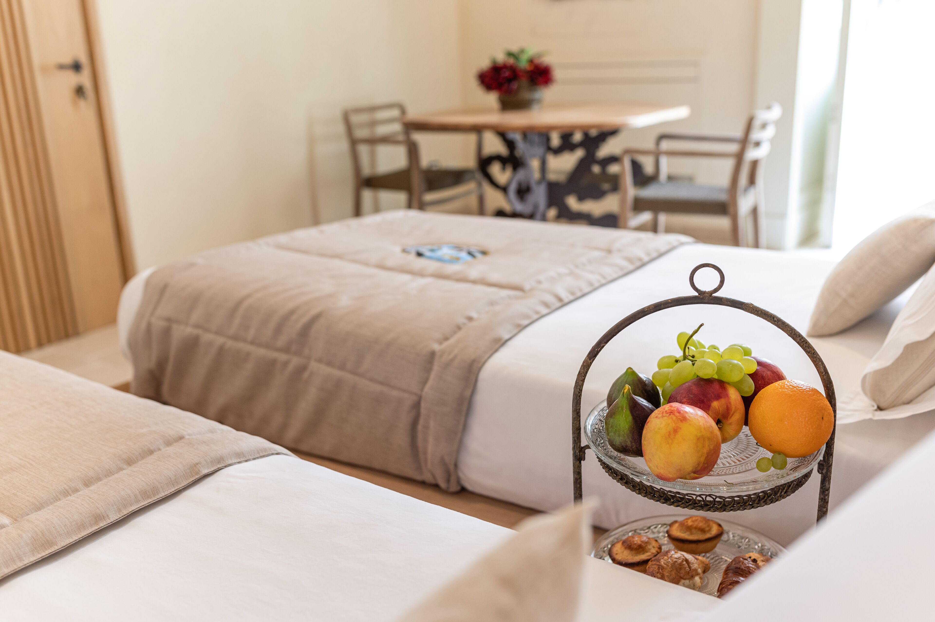 Een opgemaakt bed met een beige deken, een nachtkastje met een fruitmand, en een eethoek op de achtergrond.