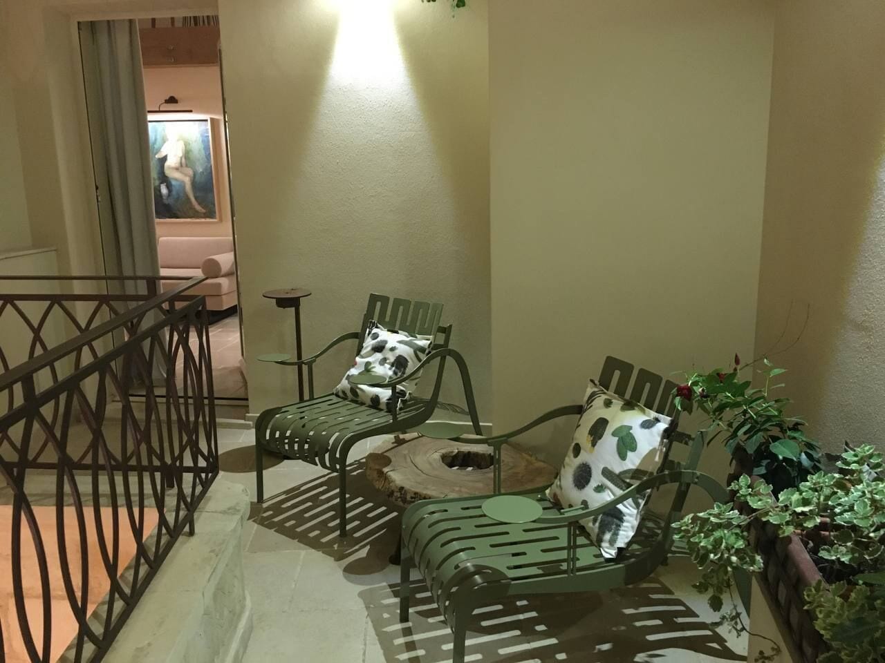 A small, cozy seating area with two green metal chairs and a round table, featuring a decorative pillow and a potted plant.