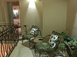 Un rincón pequeño y acogedor con dos sillas metálicas verdes y una mesa redonda, decorado con un cojín y una planta en maceta.
