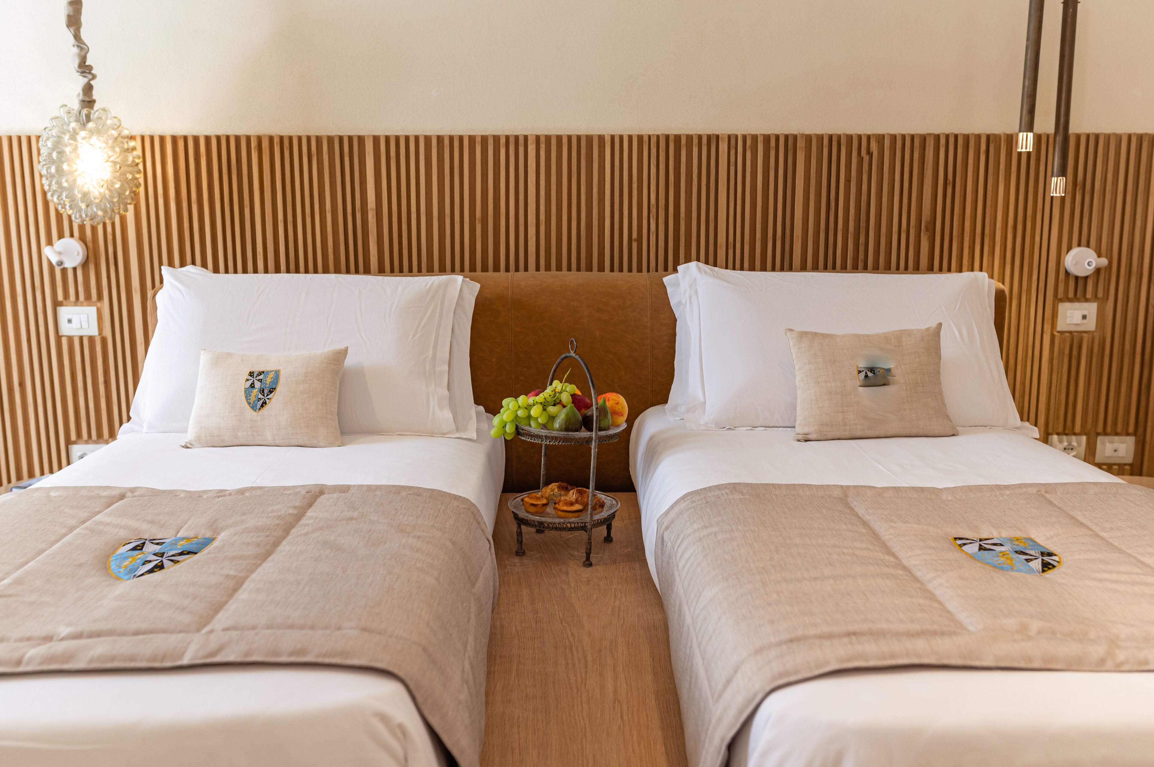 A hotel room with two beds, a wooden headboard, a small table with fruit and pastries, and a hanging light fixture.