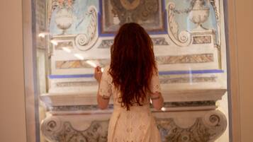 A woman in a white dress standing in front of a decorative wall with a painting and two vases.