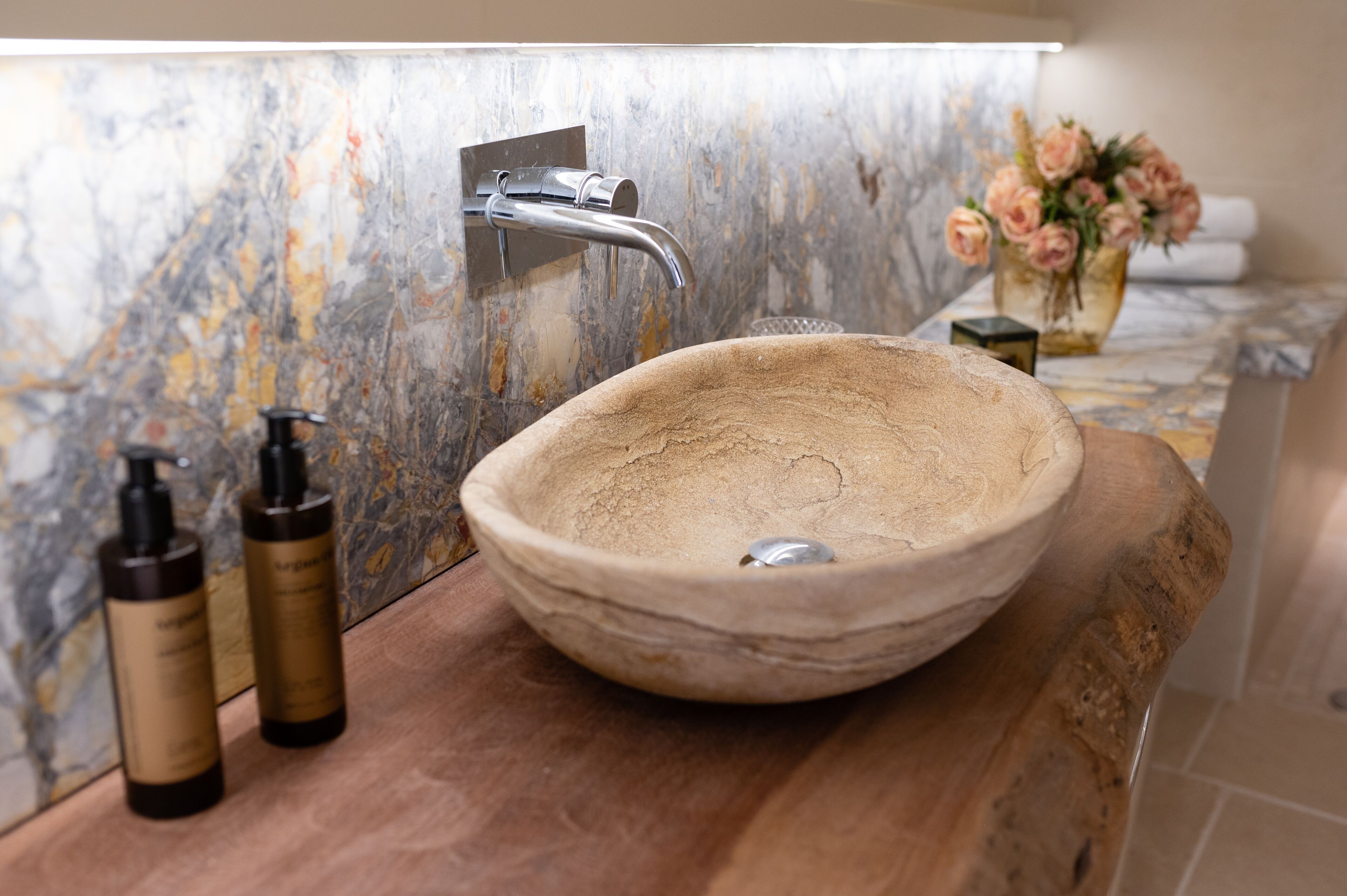 Une salle de bain avec un mur en marbre, un meuble-lavabo en bois, un lavabo rond en céramique, un robinet moderne et un vase de fleurs.