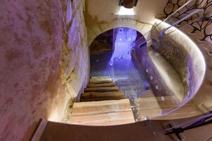 Una escalera en espiral de madera que desciende, rodeada de muros de piedra y una barandilla de metal.