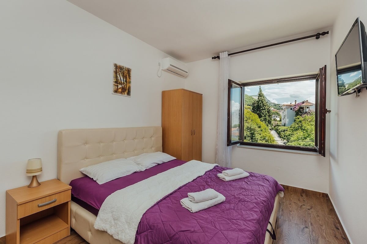 Ein Schlafzimmer mit Bett, Nachttisch, Kleiderschrank und Fenster mit Blick auf Grünflächen und Gebäude.