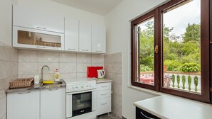 Een kleine keuken met witte kastjes, een ingebouwde oven, een spoelbak en een raam met uitzicht op groen.