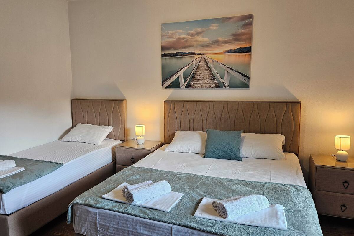 Ein Hotelzimmer mit zwei Betten, jeweils mit Kopfteil, Nachttischen und Lampen. Ein Gemälde, das einen Sonnenuntergang über einer Brücke zeigt, hängt an der Wand.