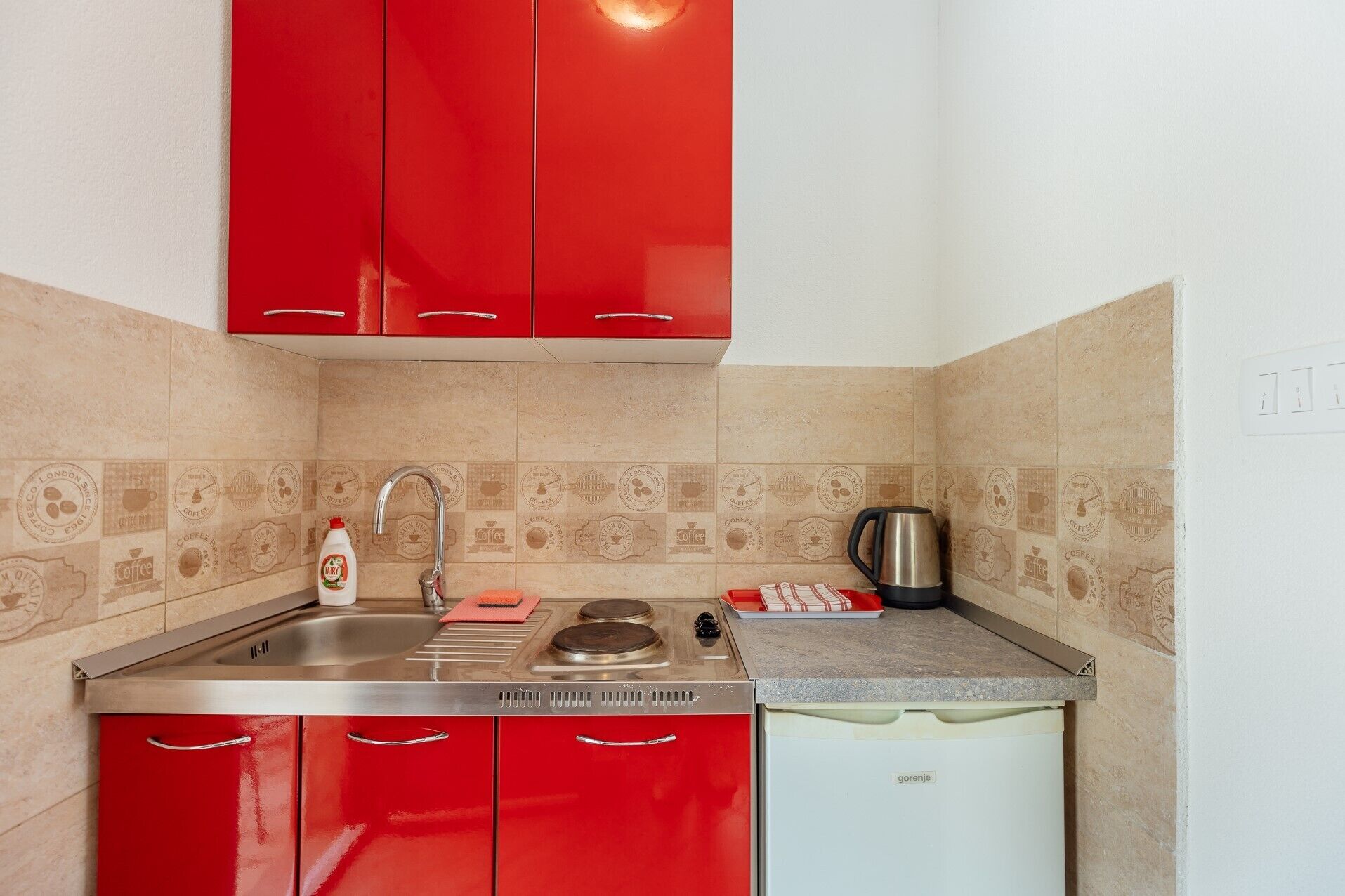 A compact kitchen with red cabinets, a stainless steel sink, a kettle, and a small refrigerator.