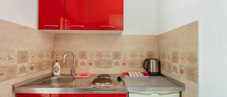 A compact kitchen with red cabinets, a stainless steel sink, a kettle, and a small refrigerator.