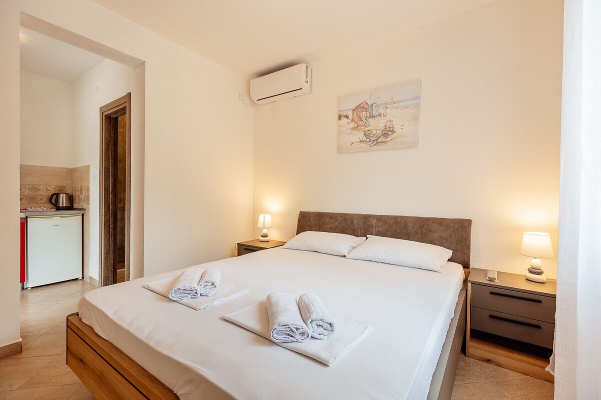 A hotel room with a bed, two bedside tables with lamps, a small kitchenette, and a painting on the wall.