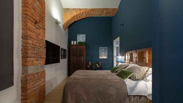 A bedroom with a brick wall, a large bed, a wooden headboard, and a mounted TV.