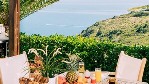 A wooden table set for breakfast with tropical fruits and beverages, overlooking a scenic ocean view. - Aries Villas | Villa Sagittarius – Private Pool & Aegean Sea Views (Skiathos)