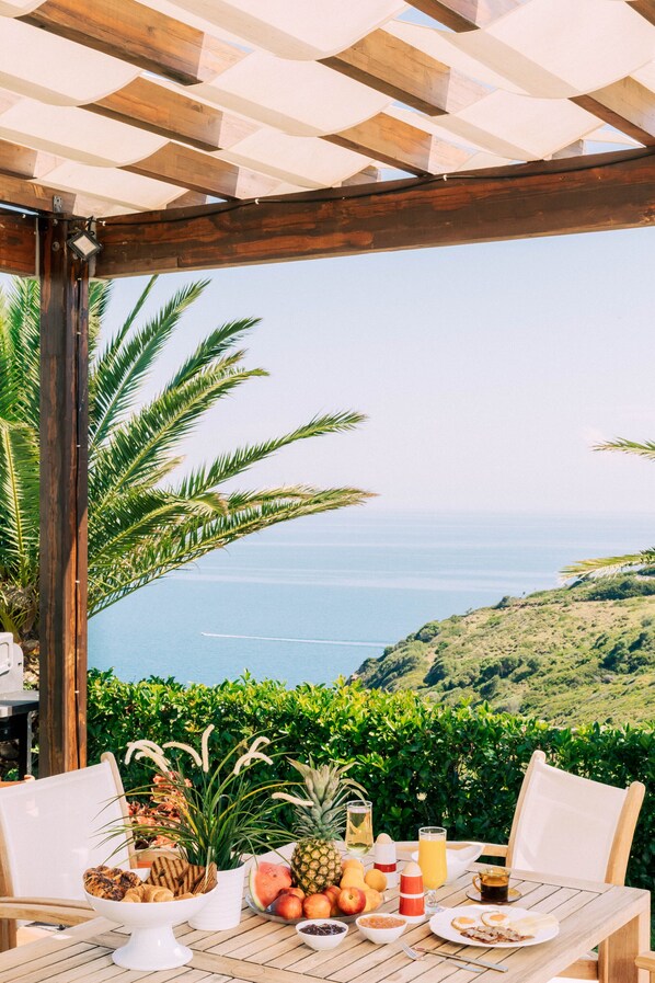 Une table en bois, dressée pour le petit-déjeuner avec des fruits tropicaux et des boissons, offrant une vue panoramique sur l’océan.