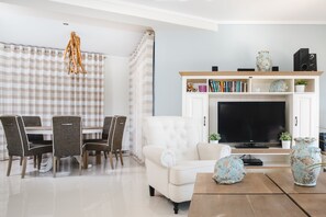 Un salon moderne avec un coin repas, comprenant un téléviseur à écran plat, des canapés blancs, une table basse en bois et une bibliothèque agrémentée d’objets décoratifs.